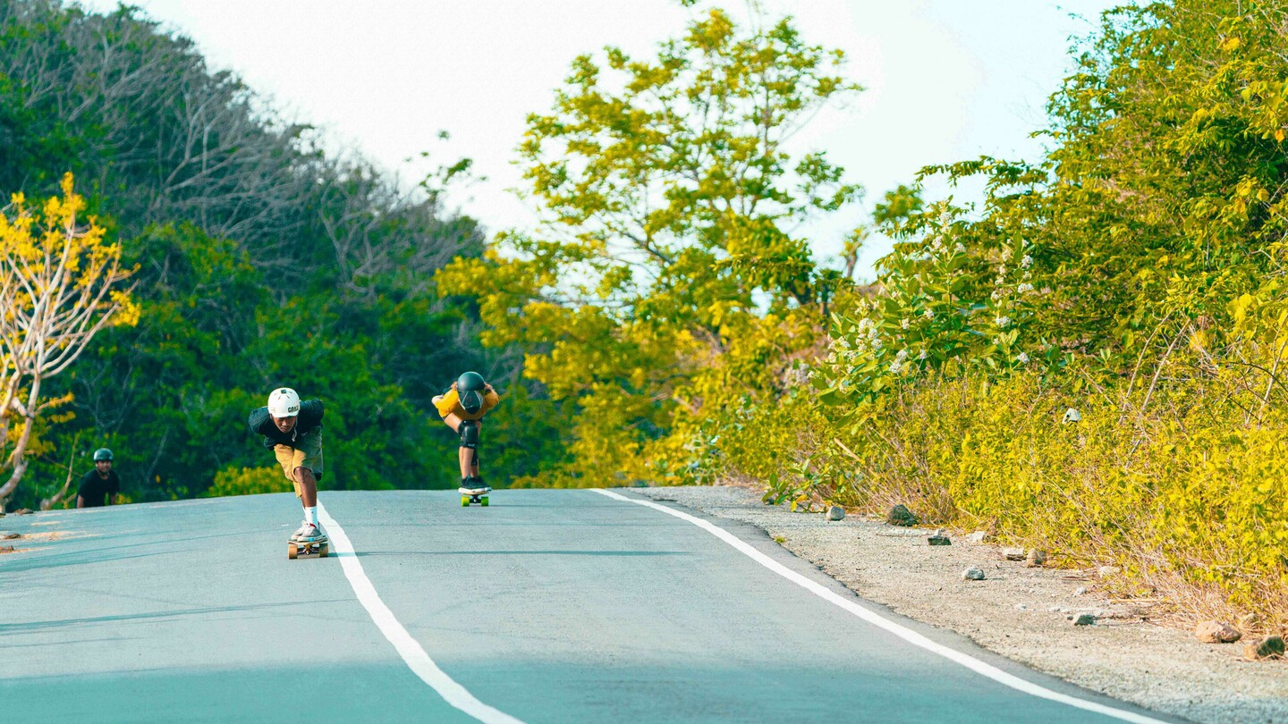 Plusieurs skateboardeurs de descente, dont Anna Pixner portant un T-shirt jaune et un casque noir, descendent une route rurale légèrement en pente. La scène est entourée de végétation luxuriante et d'arbres.