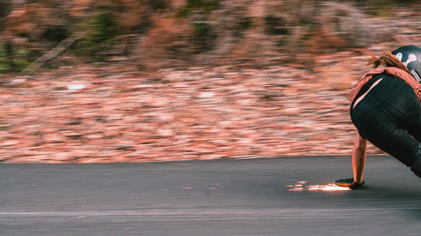 La skateboardeuse de descente autrichienne Anna Pixner est capturée en pleine vitesse sur son longboard. En position accroupie, elle touche l’asphalte avec sa main gantée, créant des étincelles. Le flou de mouvement à l’arrière-plan souligne la grande vitesse et le caractère dynamique de la scène alors qu’Anna prend un virage sur la route.