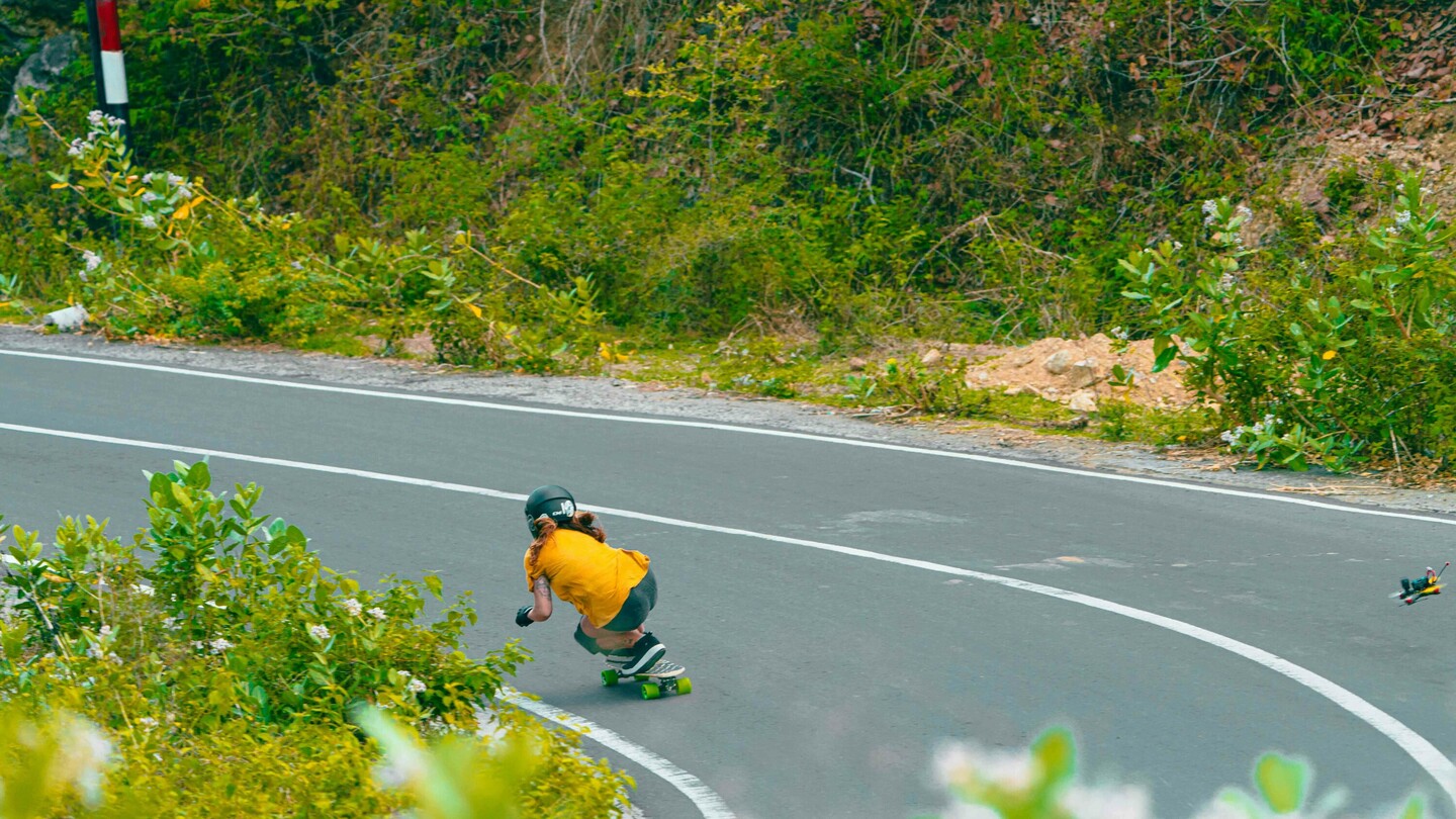 Anna Pixner, een downhill-skateboardster, rijdt in een gebogen houding door een scherpe bocht op een landelijke weg. Ze draagt een zwarte helm, een geel T-shirt en beschermende uitrusting. De omgeving is omgeven door dichte vegetatie.
