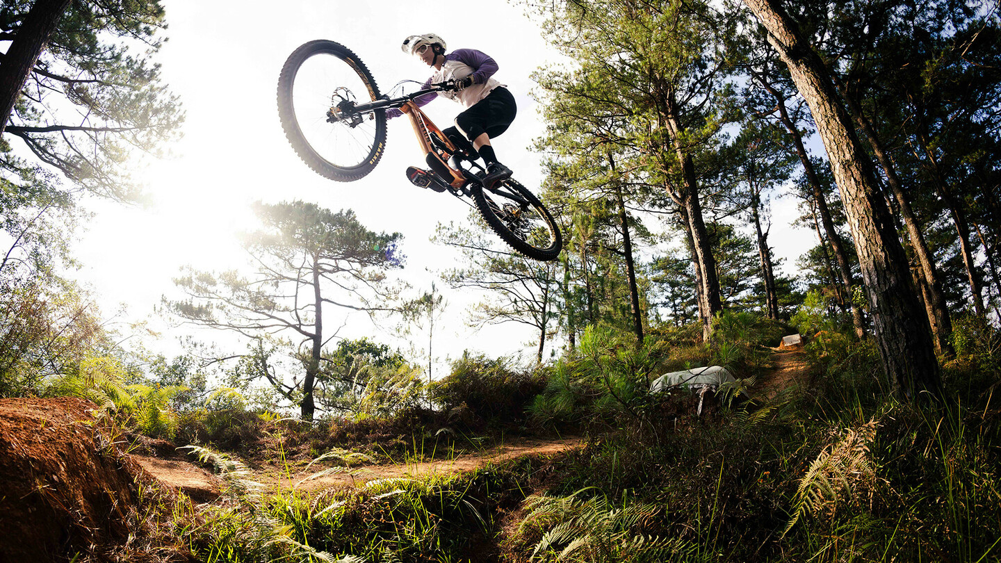 Un vététiste effectue un saut spectaculaire au-dessus d'une colline dans une zone boisée. Le cycliste porte un équipement de protection et un casque, tandis que le soleil brille à travers les arbres, baignant la forêt d'une lumière chaleureuse.