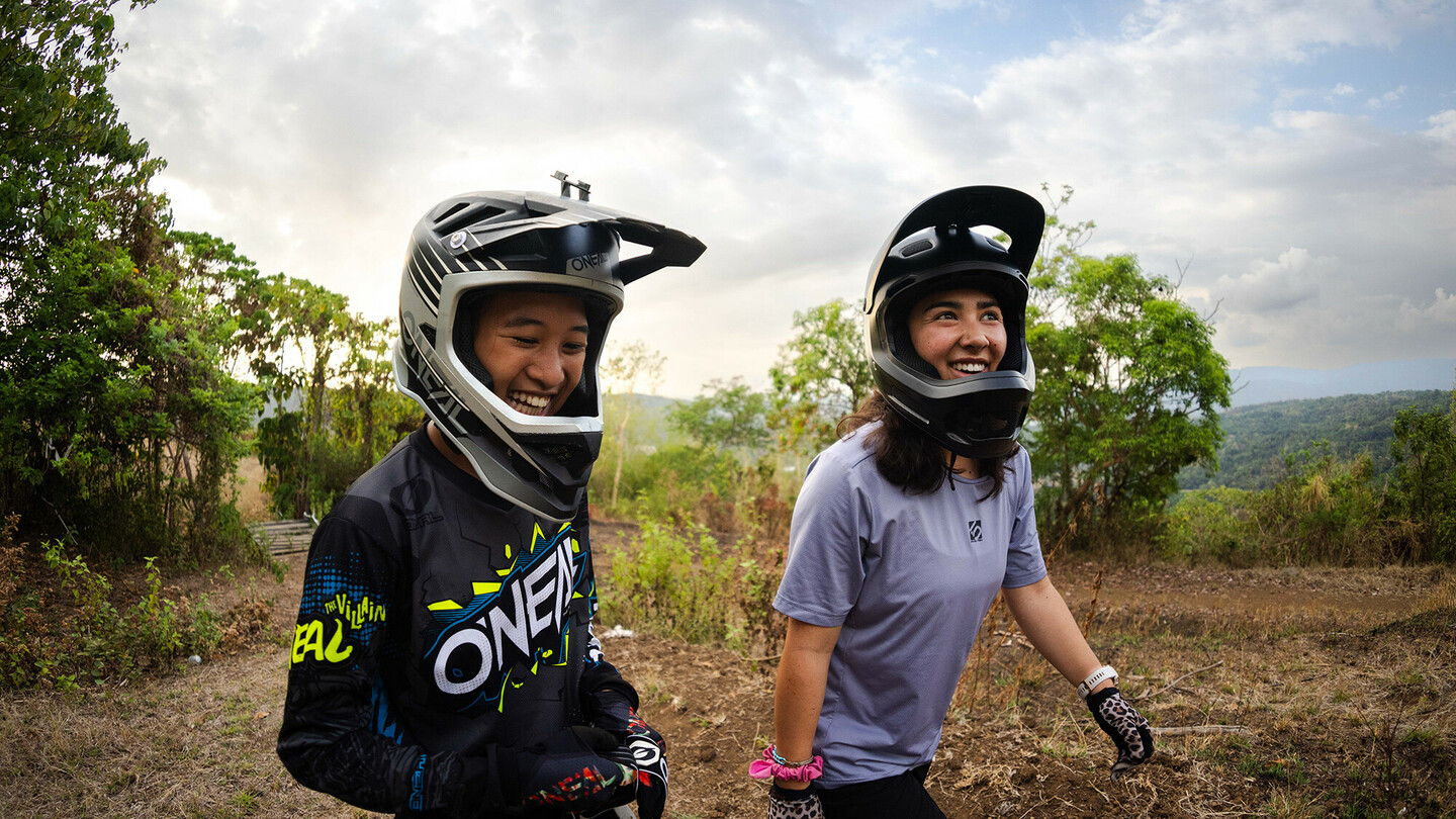 Deux jeunes femmes portant des casques intégraux sourient dans un cadre extérieur avec des arbres en arrière-plan. La personne à gauche porte un maillot foncé et celle à droite un t-shirt violet et des gants à pois.