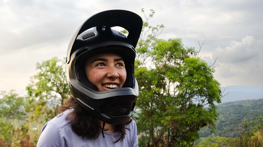 Samantha Soriano glimlachend met een zwarte fullface-helm buiten. Op de achtergrond zijn bomen en een bebost heuvelgebied te zien, met een licht bewolkte lucht.