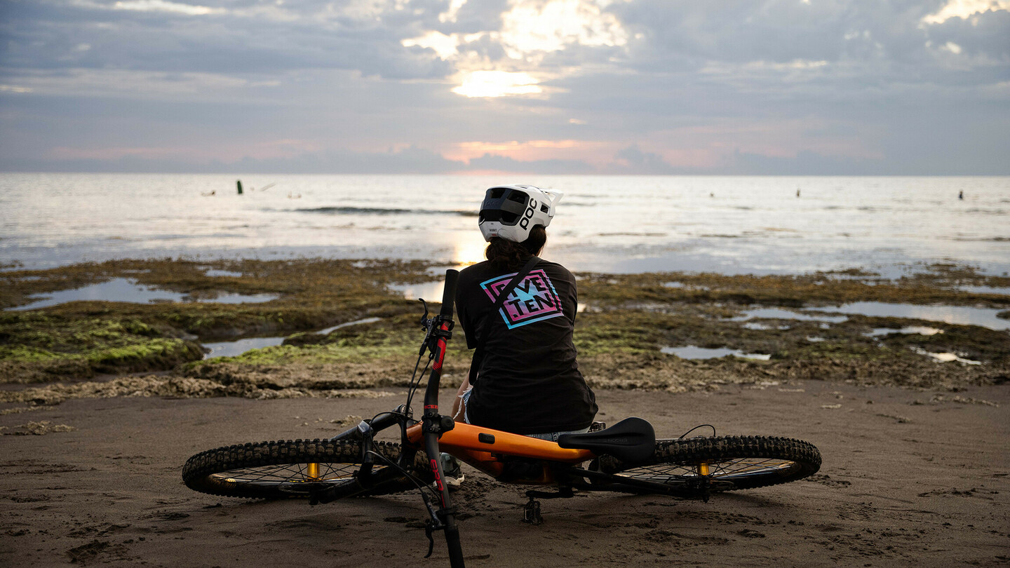 Une personne portant un casque est assise sur la plage à côté d'un VTT, regardant la mer alors que le soleil se couche derrière les nuages. L'atmosphère est calme et détendue.