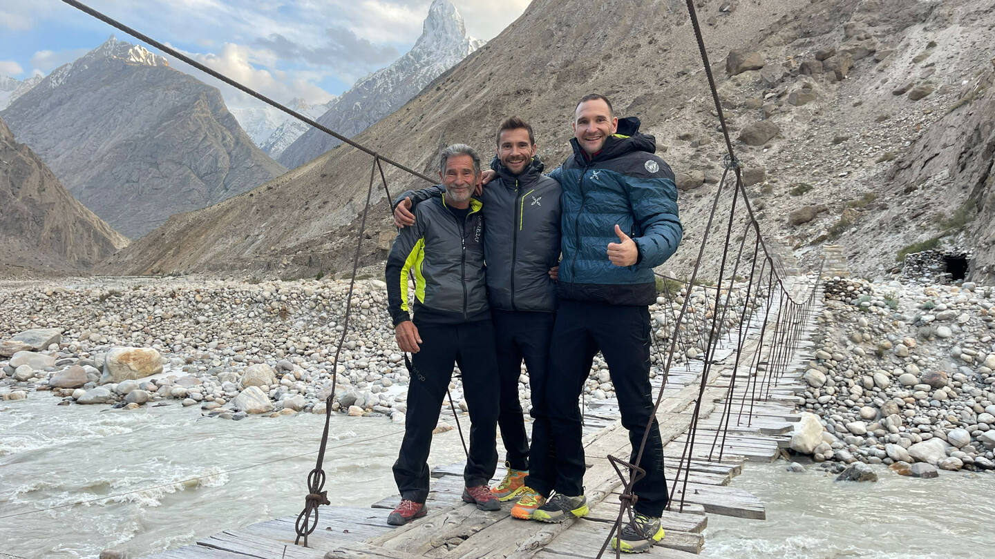 Trois alpinistes (Eduard and Alex Marin with their father) debout et souriants sur un pont suspendu au-dessus d'une rivière de montagne, entourés d'un paysage rocheux et de sommets enneigés en arrière-plan.