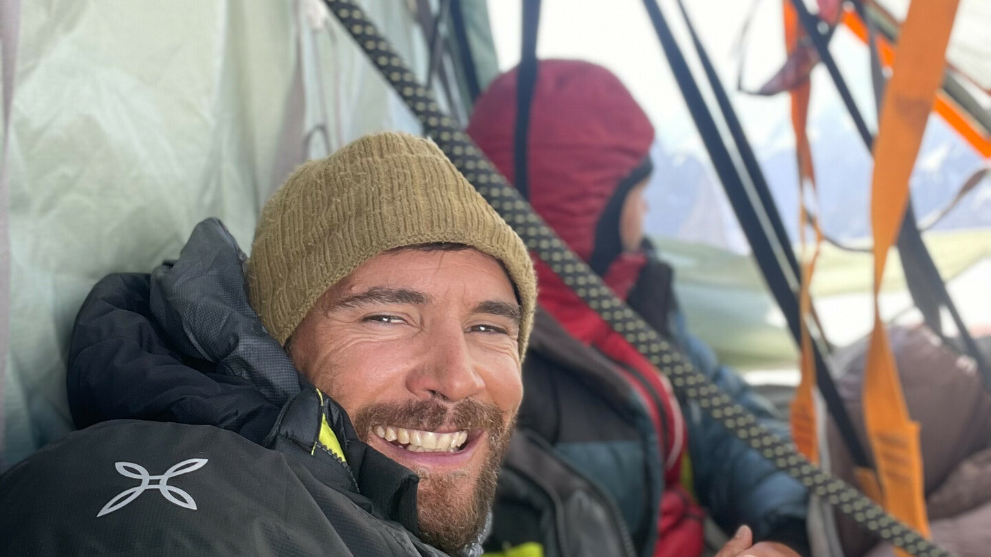 Eduard Marín souriant dans une tente lors d'une expédition en montagne, vêtu de vêtements chauds, avec un autre grimpeur en arrière-plan.