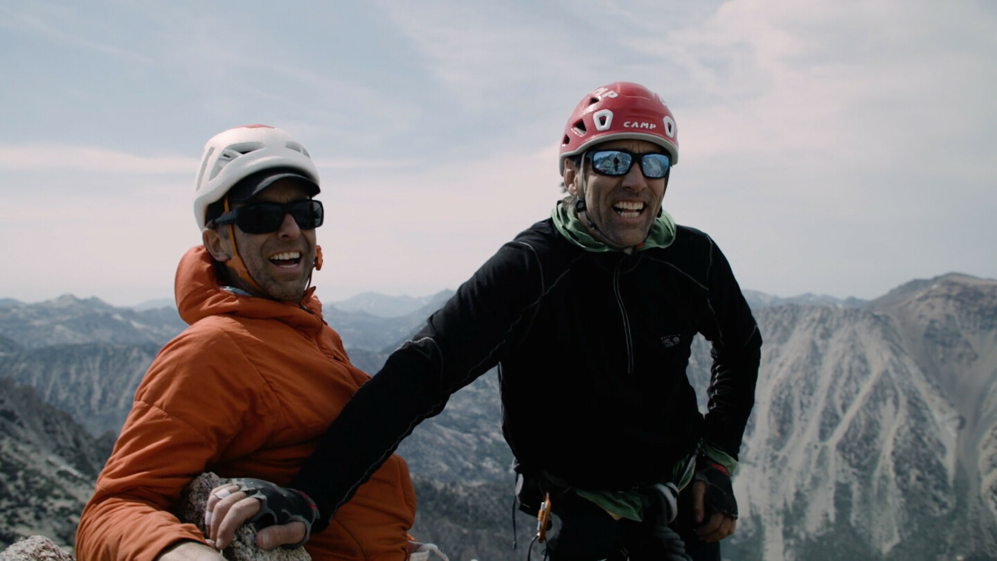 À gauche se tient Timmy O’Neill avec une veste orange et un casque, souriant avec des lunettes de soleil. À sa droite, le grimpeur aveugle Erik Weihenmayer porte une veste noire et un casque rouge. Les deux hommes se tiennent sur un sommet de montagne, heureux, profitant du vaste paysage montagneux en arrière-plan. L'image reflète l'aventure, l'escalade et la motivation.