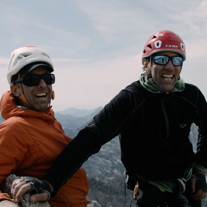 Links op de foto staat Timmy O’Neill in een oranje jas en helm, glimlachend met een zonnebril op. Rechts van hem staat de blinde bergbeklimmer Erik Weihenmayer in een zwarte jas en rode helm. Beide mannen staan op een bergtop, zien er gelukkig uit en genieten van het uitgestrekte berglandschap op de achtergrond. De afbeelding symboliseert avontuur, klimmen en doorzettingsvermogen.