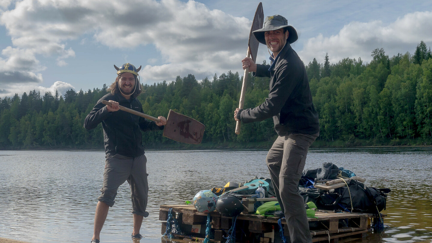 Deux hommes posent en s'amusant avec des pagaies improvisées sur la rive d'un lac. L'homme à gauche porte un casque de viking, tandis que celui à droite porte un large chapeau. Ils se tiennent à côté d'un radeau de fortune fait de palettes en bois, chargé d'équipement. En arrière-plan, on voit une rive boisée.