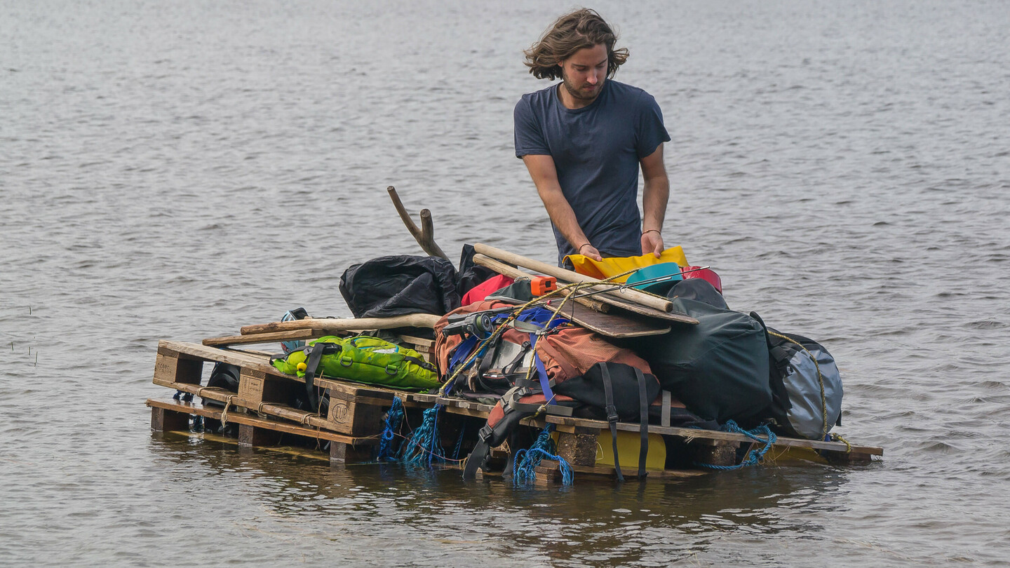 Un homme aux cheveux mi-longs se tient sur un radeau de fortune fait de palettes en bois sur un lac. Le radeau est chargé de divers objets, tels que des sacs, des pagaies et de l'équipement. En arrière-plan, on aperçoit une rive avec une forêt dense.