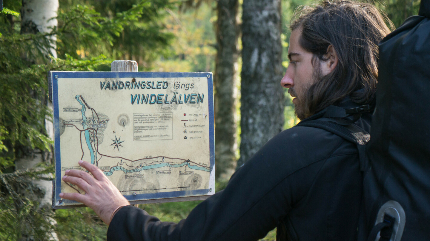 Een man met schouderlang haar en een grote rugzak bestudeert een wandelkaart met de tekst “Vandringsled längs Vindelälven” in een bos. Hij raakt de kaart aan met zijn hand en lijkt geconcentreerd. Om hem heen zijn bomen en dicht bosgebied.