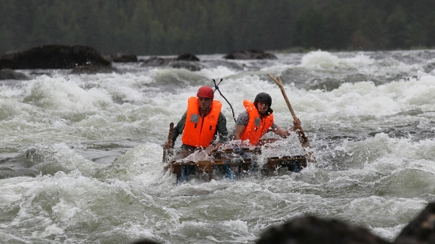 Deux personnes portant des casques et des gilets de sauvetage orange vif naviguent avec des pagaies sur un radeau de fortune à travers des rapides tumultueux et écumants.