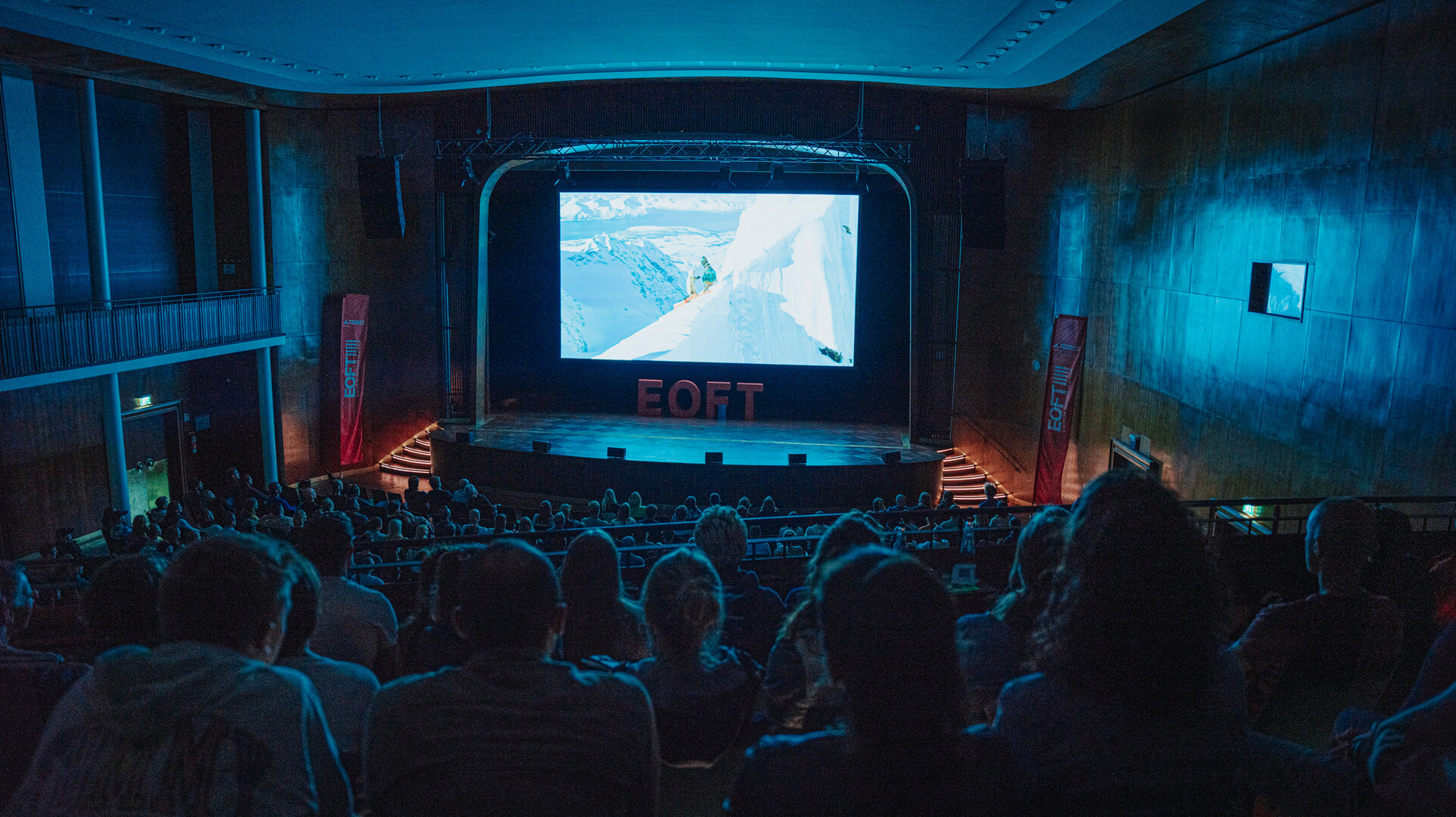 Saal mit Publikum und Leinwand, auf der eine Filmszene am eingeschneiten Berg zu sehen ist.