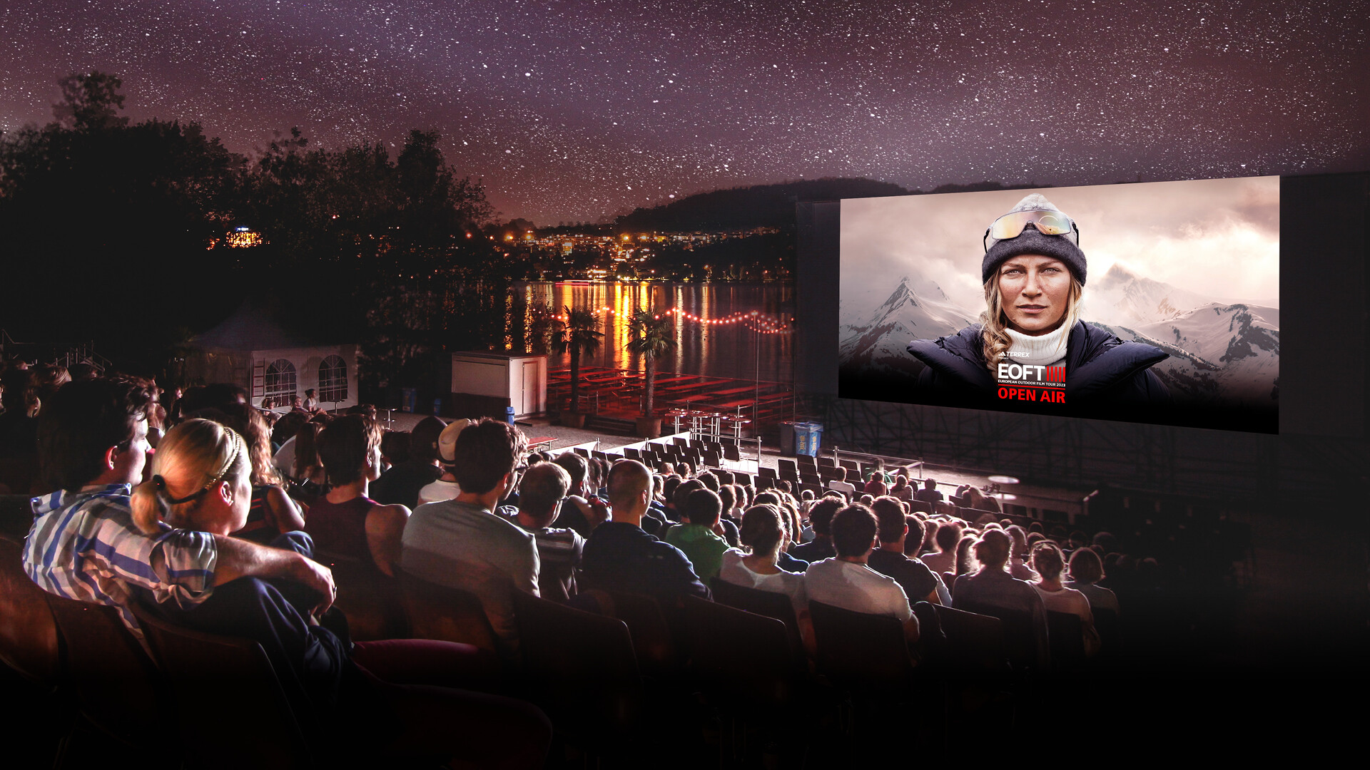 Un cinéma en plein air au bord du lac avec un large public regardant un film sous le ciel étoilé. Sur l'écran, une femme en tenue d'extérieur avec des montagnes en arrière-plan, accompagné du texte “EOFT OPEN AIR”.