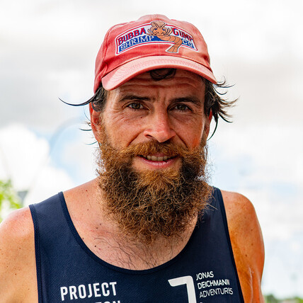 Portrait de Jonas Deichmann, un homme avec une barbe épaisse et une casquette rouge arborant le logo Bubba Gump Shrimp Co. Il porte un débardeur foncé.